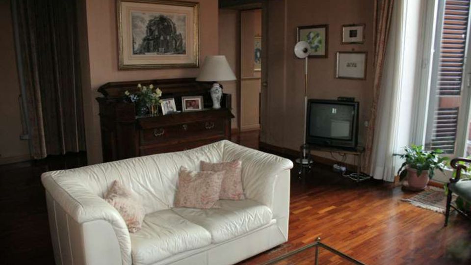 Soggiorno con pavimento in parquet e divano bianco, con cassettiera in legno e quadri alle pareti. A destra una finestra con persiane lascia entrare luce naturale; vicino ci sono un televisore e una pianta in vaso.