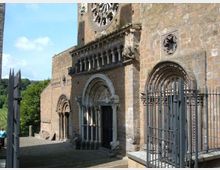 Facciata in pietra di una chiesa medievale con portali ad arco decorati, loggetta con colonnine e grande rosone sopra l’ingresso principale. Davanti si vede un sagrato in pietra con cancellate metalliche e, sullo sfondo, alberi e cielo.