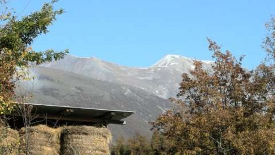 Cataste di balle di fieno coperte da una tettoia in una zona rurale recintata, circondata da alberi. Sullo sfondo si vedono montagne con cime parzialmente innevate sotto un cielo limpido.