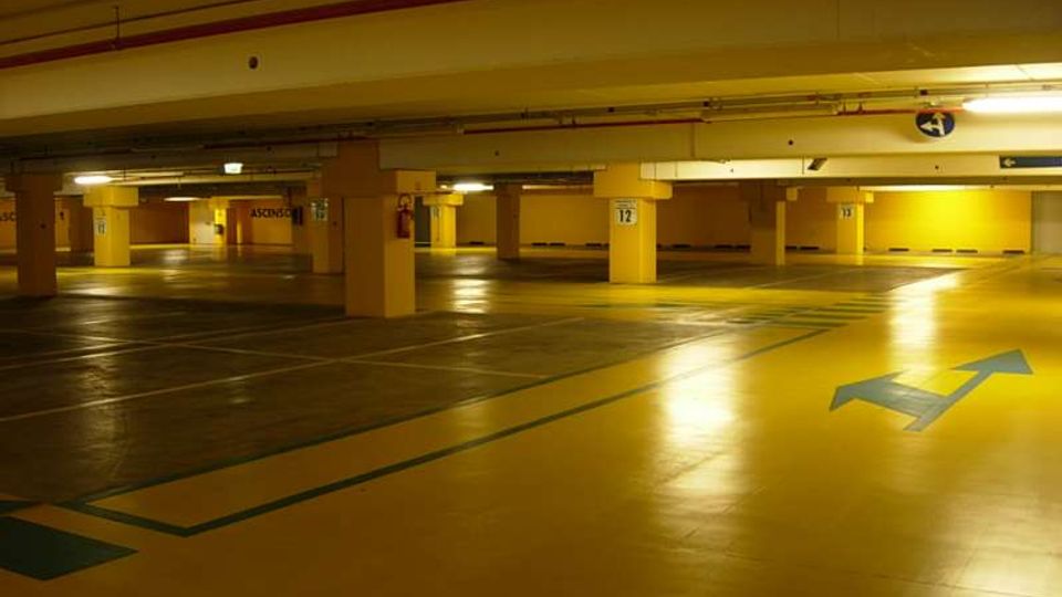 Parcheggio sotterraneo vuoto con ampie corsie asfaltate e segnaletica a terra, inclusa una grande freccia direzionale blu. Colonne in cemento numerate e illuminazione al neon sul soffitto basso con tubazioni a vista.