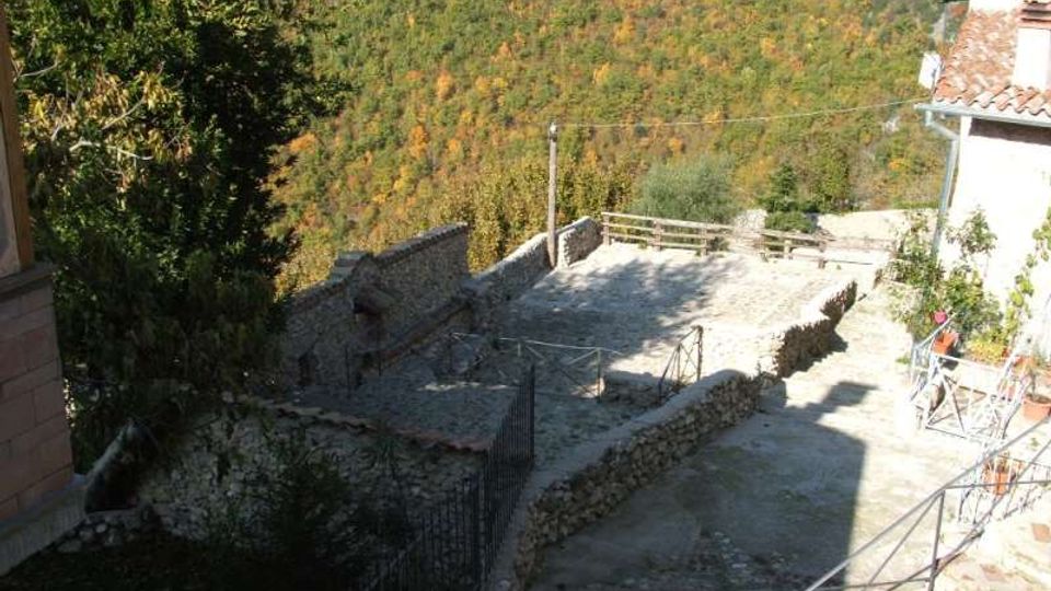 Terrazza in pietra su un pendio, delimitata da muretti e ringhiere, con pavimentazione in lastre e un piccolo spazio recintato. Sullo sfondo si vede una valle boscosa e una collina, con parte di un edificio dai tetti in coppi sulla destra.