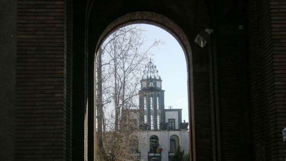 Veduta attraverso un grande arco in mattoni che incornicia un edificio con torre dell’orologio e campanile sullo sfondo. In primo piano si notano le pareti scure del portico e rami di alberi spogli contro il cielo chiaro.