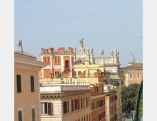 Veduta urbana con palazzi a più piani dai colori ocra e terracotta, con finestre e persiane, disposti lungo una strada. Sullo sfondo si vede un edificio monumentale con statue sul cornicione e una croce in sommità, con tetti e campanili che emergono dietro.
