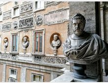 Facciata interna di un edificio storico con decorazioni in stucco e bassorilievi, finestre rettangolari e nicchie con busti scultorei. In primo piano, un busto marmoreo su piedistallo lungo un corridoio o loggia in pietra.