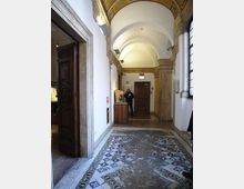 Corridoio interno con soffitto a volte e archi in muratura, pareti bianche e pavimento in mosaico decorato. Ai lati si vedono porte in legno scuro e una grande finestra, con un banco espositivo vicino al fondo.