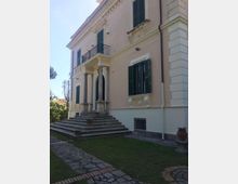 Facciata di una villa storica a due piani con intonaco chiaro, persiane verdi e cornicioni decorativi. Un ampio scalone conduce a un ingresso con portico sorretto da colonne e balconcino, affacciato su un prato con vialetto in pietra.