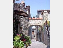 Vicolo lastricato in pietra tra edifici in muratura, con arco in pietra che collega le case e una scalinata laterale con ringhiera. Ai lati si vedono finestre, persiane e vasi di fiori, in un contesto di borgo storico all’aperto.