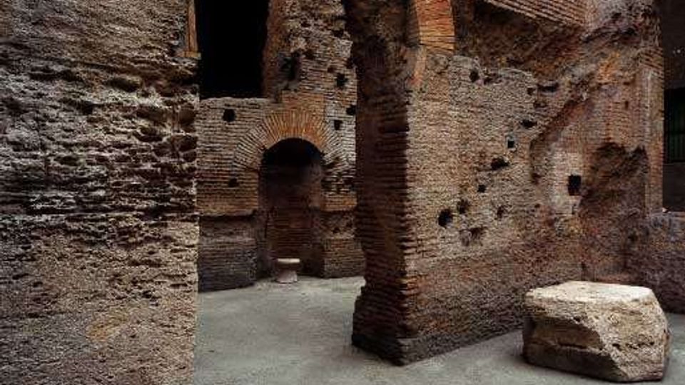 Interno di rovine in mattoni con grandi archi a tutto sesto e pareti irregolari segnate da fori e tracce di muratura antica. Sul pavimento in cemento si vede un grande blocco di pietra squadrato accanto a un corridoio che conduce ad altri passaggi ad arco.