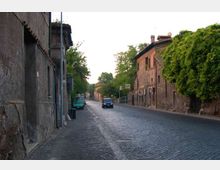Strada urbana in pavé con marciapiede stretto, fiancheggiata da edifici in muratura dall’aspetto storico. Sul lato destro si vede un edificio con finestre e alberi che coprono parte della facciata, con alcune auto lungo la carreggiata.