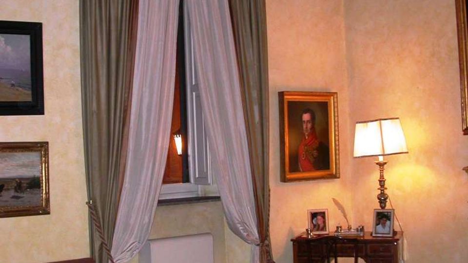Interno di una sala in stile classico con pareti chiare, pavimento in legno e finestra alta con tende drappeggiate. Accanto alla finestra ci sono un piccolo scrittoio con lampada da tavolo e cornici, quadri appesi alle pareti, un divano e tappeti decorati.