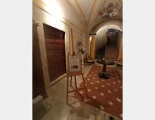 Ingresso interno di un edificio storico con pavimento in marmo e tappeto di piastrelle rosse a rombi, porta in legno imbottita e cavalletto con cartello informativo. Sul fondo un arco affrescato con stemma conduce a una scalinata, con tavolino rotondo, sedie e lampadario.