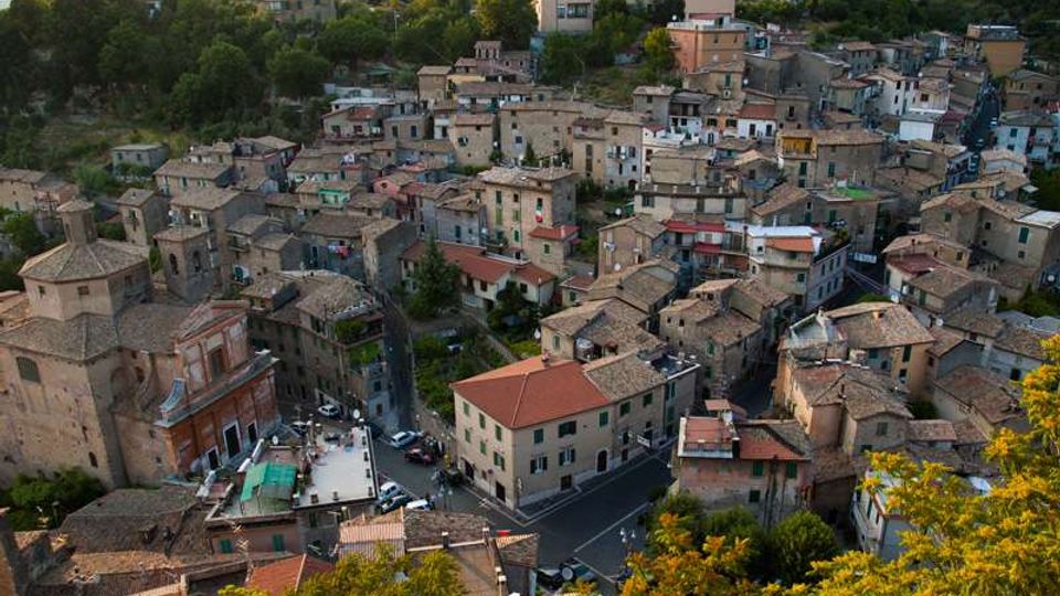 Veduta dall’alto di un borgo collinare con case in pietra addossate tra loro e tetti in tegole, attraversato da strade strette che convergono in un piccolo incrocio. Intorno si vedono alberi e vegetazione sui pendii, con alcune abitazioni più recenti sullo sfondo.