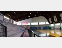 Interno di un palazzetto dello sport con parquet segnato per basket e pallamano, spalti in cemento su un lato e ringhiera lungo il corridoio. Tetto alto ad arco con travi a vista, grandi finestre e pareti con pannelli e striscioni.