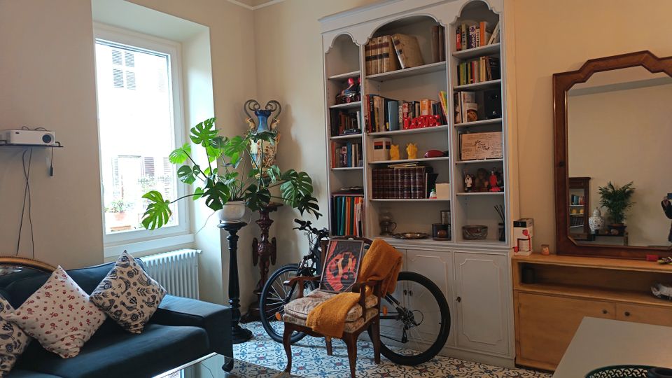 Soggiorno luminoso con grande finestra, pavimento a piastrelle decorate e divano scuro con cuscini. Sulla parete c’è una libreria bianca piena di libri e oggetti, con una bicicletta appoggiata davanti e, a lato, un comò in legno con grande specchio.