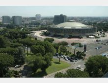 Vista aerea di un grande edificio moderno a pianta circolare con cupola bassa e facciata vetrata, circondato da strade e parcheggi. Intorno si estendono aree verdi con alberi e, sullo sfondo, palazzi e grattacieli della città.