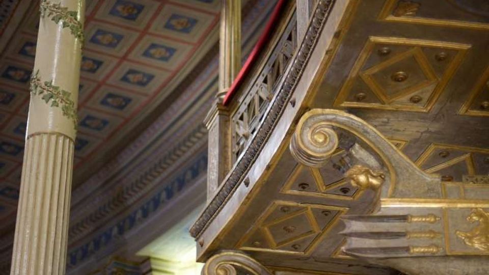 Interno di un edificio storico con colonne alte decorate e un balcone curvo con balaustra in ferro; il soffitto a cassettoni è dipinto con motivi geometrici e dettagli dorati. In primo piano si vedono mensole scolpite con volute e ornamenti che sorreggono la galleria.