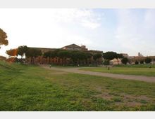 Ampio prato urbano con sentieri sterrati e filari di pini marittimi, con un lungo edificio storico in mattoni sullo sfondo. Cielo aperto e spazi verdi estesi che definiscono un’area di parco o piazza.