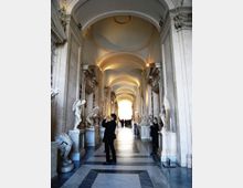 Lungo corridoio monumentale in pietra con alte arcate e soffitto a volte illuminato da luci calde. Ai lati si trovano nicchie e piedistalli con statue classiche in marmo e pavimento a motivi geometrici che conduce verso un’uscita luminosa in fondo.