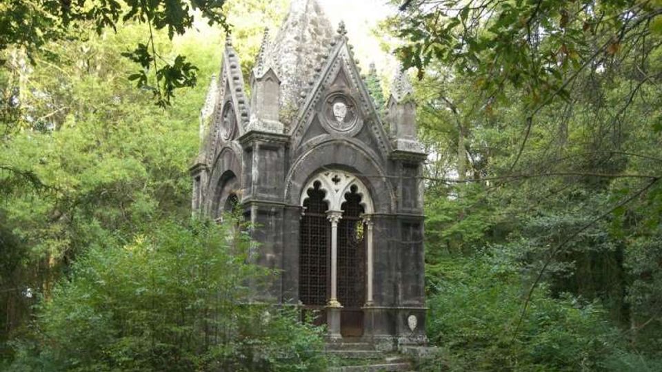 Piccola cappella in pietra in stile gotico, con archi a sesto acuto, finestre traforate e cuspidi decorative, situata in un’area boschiva. Vegetazione fitta e rami degli alberi circondano l’edificio e una breve scalinata conduce all’ingresso.