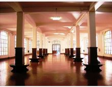 Ampio spazio interno tipo capannone o sala industriale con pavimento lucido, file di colonne quadrate con basamenti scuri e soffitto alto con lampade rettangolari. Lungo le pareti ci sono grandi finestre a riquadri e in fondo una porta vetrata che dà accesso all’esterno.