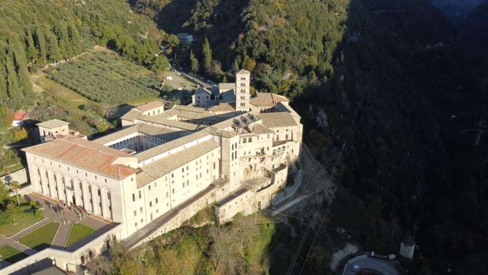 Veduta aerea di un grande complesso monastico in pietra chiara con tetti in coppi rossi, chiostro interno e campanile, costruito sul bordo di una scarpata. L’edificio è circondato da boschi e campi coltivati, con strade e piccoli edifici rurali nelle vicinanze.