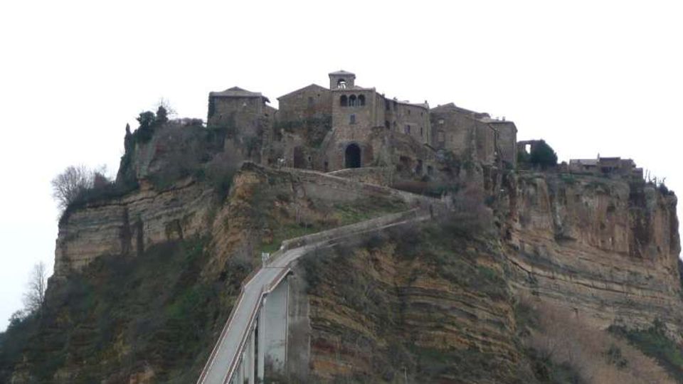 Borgo storico in pietra arroccato sulla sommità di una rupe di tufo stratificato, con edifici compatti e una chiesa con piccolo campanile. Una lunga strada sopraelevata con parapetti sale dal basso fino all’ingresso del paese lungo il crinale della scogliera.