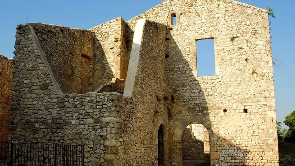 Ruderi di un edificio in pietra con muri alti e aperture senza infissi, tra cui una grande finestra rettangolare e un arco di passaggio al piano terra. La struttura è priva di tetto e si trova all’aperto sotto un cielo sereno.