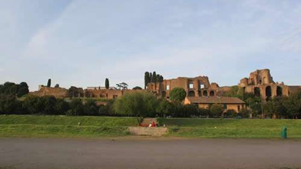 Panorama di rovine archeologiche in mattoni su una leggera altura, con archi e murature antiche tra alberi alti e vegetazione. In primo piano si vede un ampio prato con un sentiero asfaltato e cielo sereno sullo sfondo.
