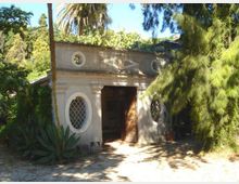 Piccolo edificio in muratura in un giardino, con facciata classica decorata, ingresso centrale con porta in legno e due finestre ovali con griglie. Intorno ci sono alberi alti, palme e vegetazione fitta che ombreggia il vialetto d’accesso.