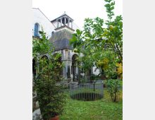 Cortile-giardino con prato e alberi di agrumi, con un pozzo circolare recintato in metallo al centro. Sullo sfondo si vede un edificio storico in pietra con portici ad archi e un piccolo campanile sopra un tetto a padiglione.