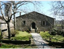 Edificio in pietra a un piano con ingresso ad arco raggiunto da una breve scalinata, affacciato su un giardino con prato e un vialetto centrale. Alberi spogli e vasi in terracotta circondano il percorso, con altri edifici in pietra sullo sfondo.