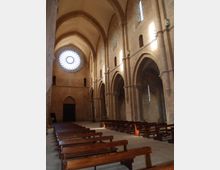Interno di una chiesa in pietra con navata ampia, volte a ogiva e arcate laterali sorrette da colonne. In fondo si vede un grande rosone circolare e file di panche in legno lungo il pavimento in pietra.