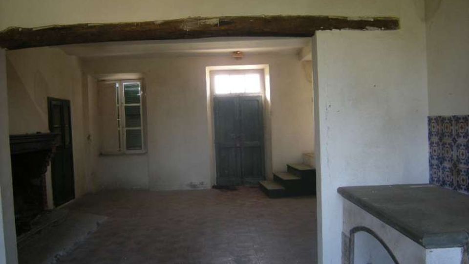 Interno rustico e spoglio con pavimento in cotto, pareti intonacate e una grossa trave in legno a vista che attraversa la stanza. Sul fondo ci sono una porta in legno rialzata da alcuni gradini e una finestra, con un piano in muratura sul lato destro e una sezione di piastrelle decorative.