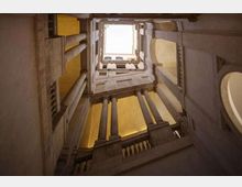 Veduta dal basso all’interno di una corte verticale in pietra, con colonne e balaustre su più livelli, che conduce a un lucernario quadrato aperto verso il cielo. Le pareti presentano archi e aperture laterali illuminate da luce calda.