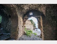 Interno in pietra di una struttura antica con volta ad arco, che inquadra un’apertura irregolare verso l’esterno. Attraverso il varco si vedono mura in rovina, vegetazione e una spiaggia rocciosa sul mare.