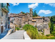 Borgo storico in pietra su uno sperone roccioso, con case addossate e una torre campanaria che domina il centro abitato. In primo piano una passerella con ringhiera e scalinate conduce verso il paese, affiancata da vegetazione e alberi sotto un cielo azzurro.