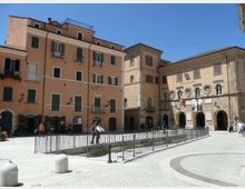 Piazza urbana lastricata con area centrale recintata da ringhiere metalliche, circondata da edifici storici a più piani con facciate color ocra e finestre con persiane. Sul lato destro si vede un palazzo con portici ad archi e stemmi sulla facciata, con bandiere esposte.