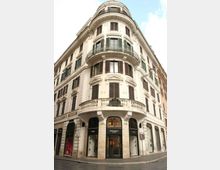Edificio storico d’angolo in stile neoclassico, con facciata chiara, finestre con persiane scure e balconi curvi con ringhiere in ferro battuto. Al piano terra si trovano vetrine di negozi e ingressi su una strada urbana stretta.