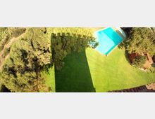 Vista dall’alto di un’area verde con prato curato, circondata da alberi, con una piscina rettangolare azzurra nell’angolo superiore destro. Una grande ombra di un edificio si estende sul prato e si intravede un piccolo tetto in tegole sul lato destro.