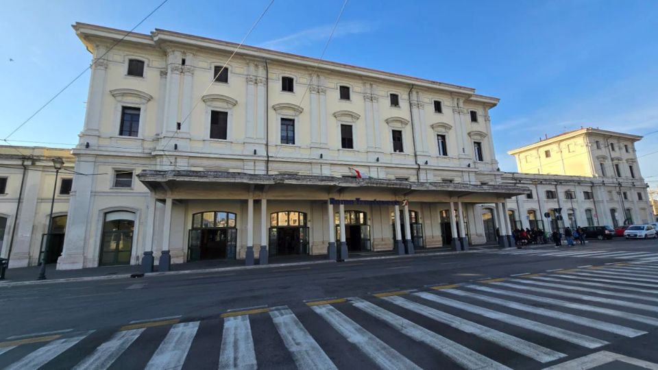 Facciata di una stazione ferroviaria storica a tre piani, con intonaco chiaro, finestre alte e un lungo portico con colonne e archi d’ingresso. Davanti si vede una strada con attraversamento pedonale a strisce e cavi aerei sopra la carreggiata.