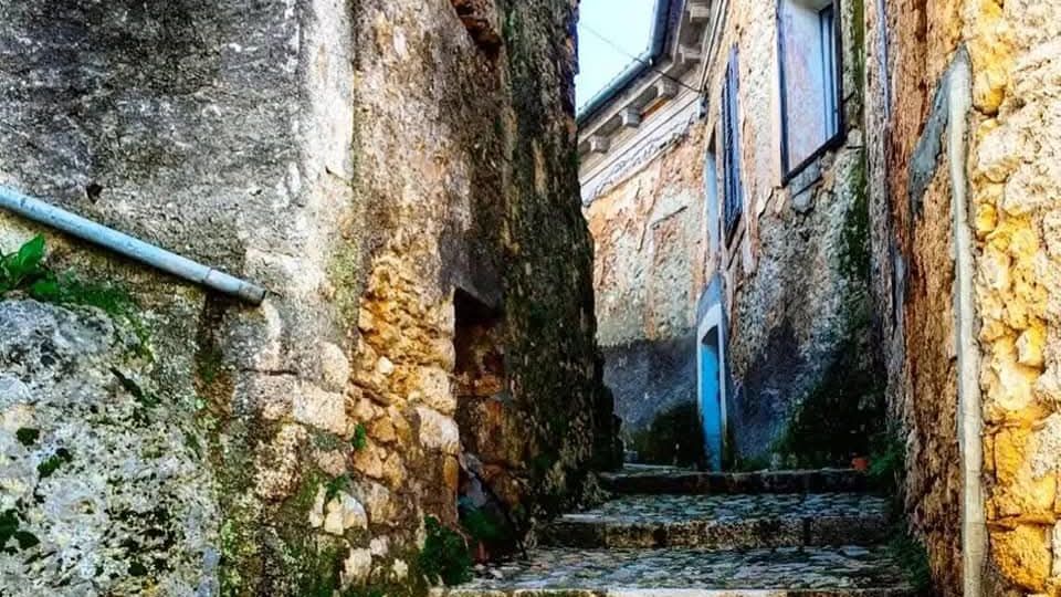 Vicolo stretto in pietra con pavimentazione acciottolata e scalini che salgono tra antiche case in muratura. Le pareti in pietra grezza mostrano segni di umidità e muschio, con finestre e una grondaia visibili lungo la salita.