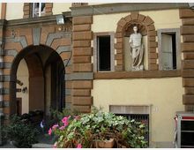 Facciata di un edificio storico con grandi blocchi bugnati in pietra e un ampio arco d’ingresso che conduce a un passaggio coperto. Al piano superiore, una statua in una nicchia arcuata è affiancata da finestre incorniciate in pietra, con vegetazione e fiori in primo piano.
