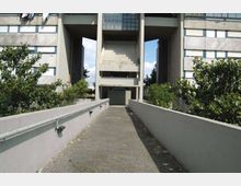 Passerella pedonale in cemento con corrimano che conduce all’ingresso di un grande edificio moderno in calcestruzzo, con ampie aperture centrali e finestre a griglia. Vegetazione e alberi fiancheggiano il percorso ai lati.