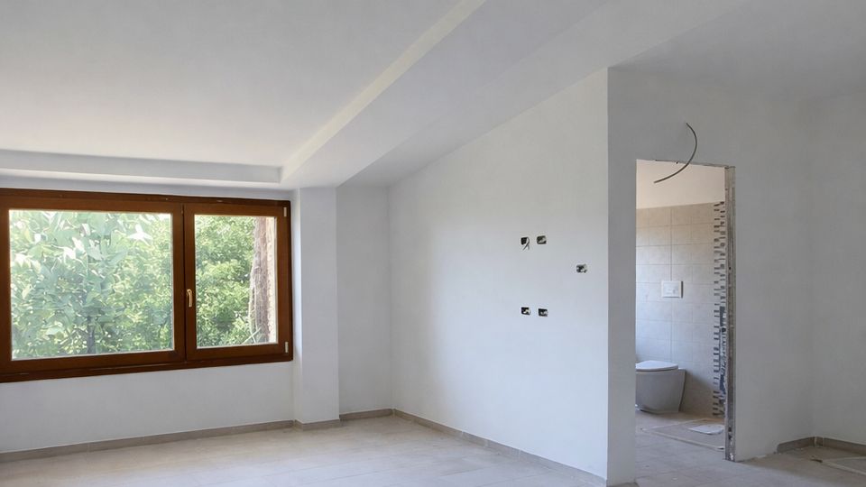 Stanza interna non arredata con pareti bianche e pavimento in piastrelle chiare, illuminata da una grande finestra con infisso in legno e vista sul verde esterno. A destra si vede un’apertura verso un bagno piastrellato, con impianti elettrici a vista e soffitto inclinato.