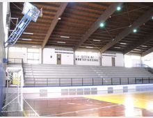 Interno di un palazzetto dello sport con campo da basket in parquet e canestri, affacciato su grandi gradinate in cemento con corrimano. Il soffitto è alto con travi in legno e luci sospese, e sulla parete di fondo compare la scritta “Città di Montefiascone”.