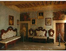 Interno di una sala storica con soffitto a travi di legno decorato, pareti rivestite di carta da parati e numerosi quadri e affreschi. Lungo le pareti sono disposti divani e poltrone imbottite in stile antico, con una vetrina in legno e una corda di delimitazione per i visitatori.