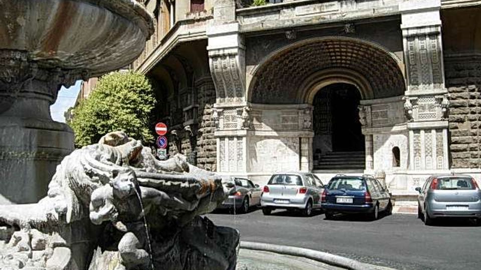 Fontana in pietra con sculture e vasca circolare in primo piano, in una piazza urbana. Sullo sfondo si vede un grande edificio storico in pietra con portico ad archi, colonne e un ingresso monumentale, con auto parcheggiate lungo la strada.