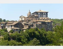 Veduta di un borgo collinare con case in pietra dai tetti in coppi, raccolte attorno a una chiesa con cupola e campanile e a un edificio più grande in posizione dominante. In primo piano si vede vegetazione fitta, con colline boscose sullo sfondo sotto un cielo sereno.