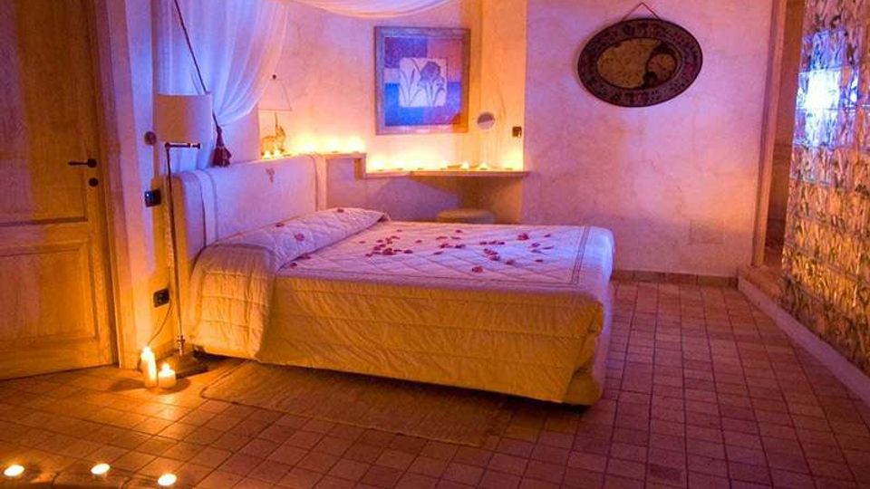 Camera da letto in stile romantico con grande letto matrimoniale, baldacchino in tessuto bianco e illuminazione soffusa a candele. Pavimento piastrellato con gradino curvo, porte in legno e parete decorativa in vetrocemento con luce integrata.