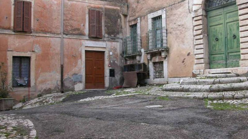 Piccola piazza in pendenza con pavimentazione in asfalto e ciottoli, circondata da edifici in intonaco rosato deteriorato con finestre a persiane e grate. A destra si vede un grande portone verde ad arco con gradini in pietra e cornici in bugnato.